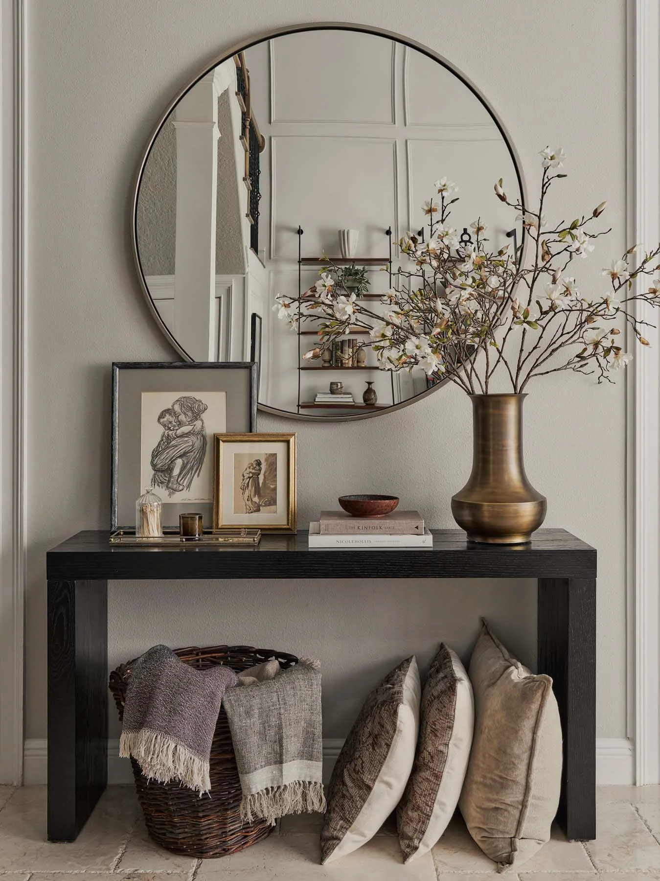A console table with framed artwork, books, a vase, and a bowl, decorated with a large vase of blossoming branches, placed against a wall with a round mirror reflecting a staircase and bookshelf.