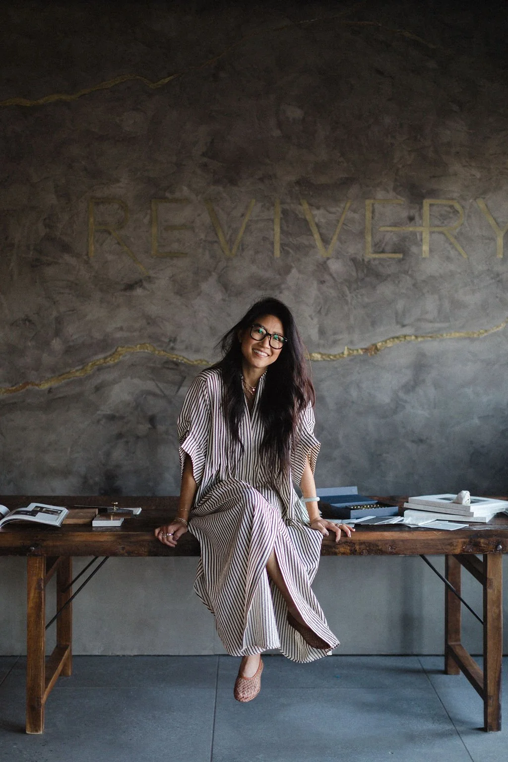 A woman with long dark hair, glasses, and a striped dress sits on a wooden table inside a room with a gray textured wall. The wall has the word 'REVIVERY' written in large gold letters.