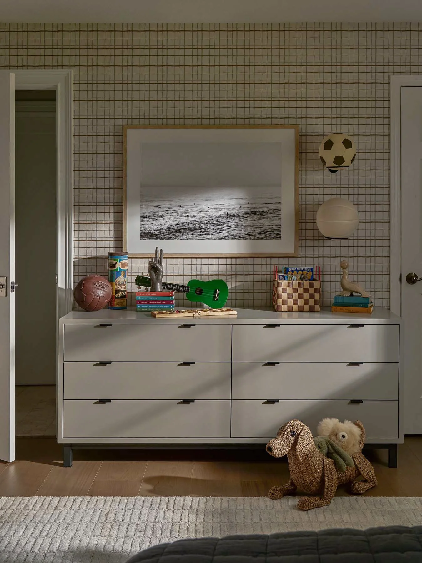 A white dresser with nine drawers against a wall with checkered wallpaper, decorated with toys including a pink soccer ball, a small green ukulele, a silver hand model, books, a basket with various items, a small dinosaur model, and stacked books. A 