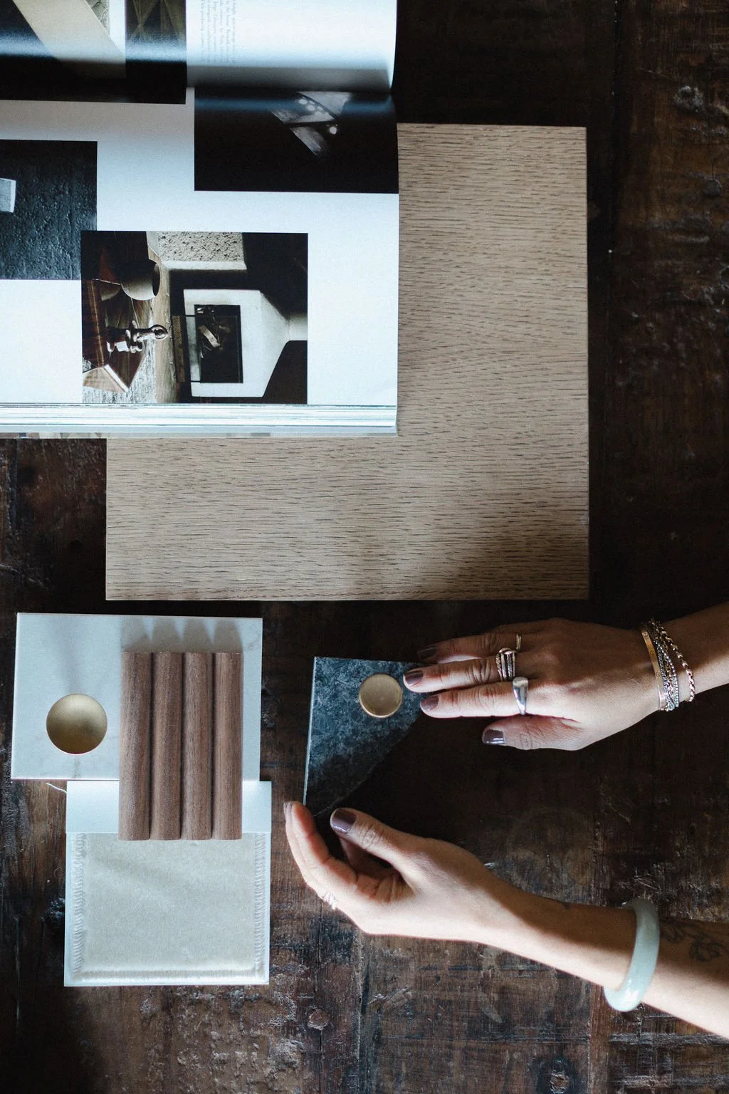 A person with rings and bracelets on their hands is holding a small sample of dark green marble with a round hole. Various material samples and a photo collage are arranged on a dark wooden table.