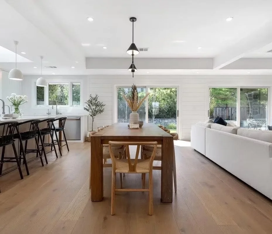 Open-concept living and dining area with white walls, light wooden floors, a large wooden dining table with chairs, a white sofa, and sliding glass doors leading outside.