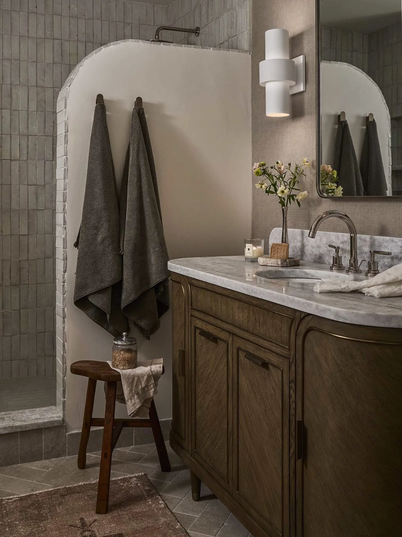 A bathroom with a wooden vanity cabinet, a marble countertop, a sink with a modern faucet, a large mirror, a white wall sconce, and a decorative vase with flowers. There are two gray towels hanging on hooks beside the shower area and a small wooden s