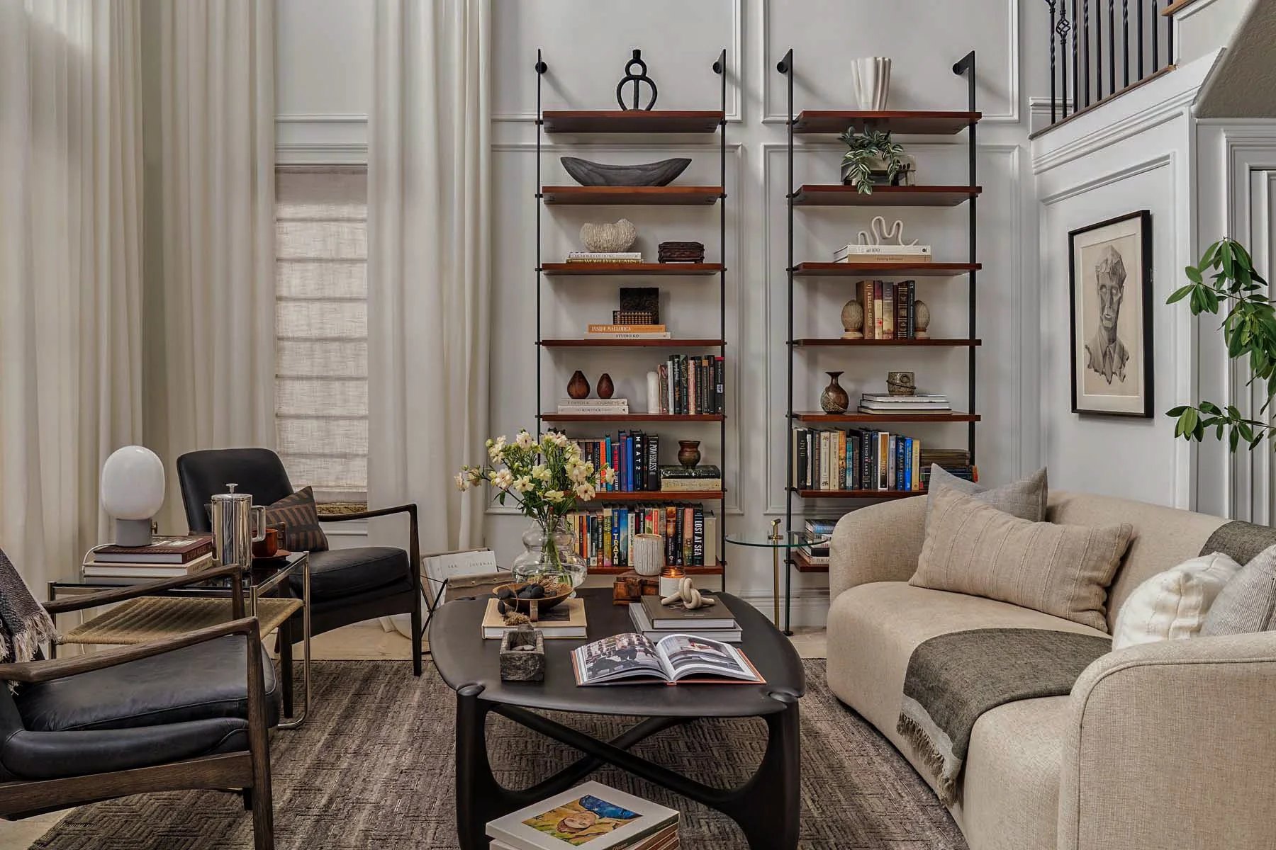 A cozy living room with bookshelves filled with books and decorative items, a beige sofa with pillows, a black armchair, a coffee table with magazines, a glass side table, and a wall art featuring a pencil sketch, with cream curtains and a potted pla