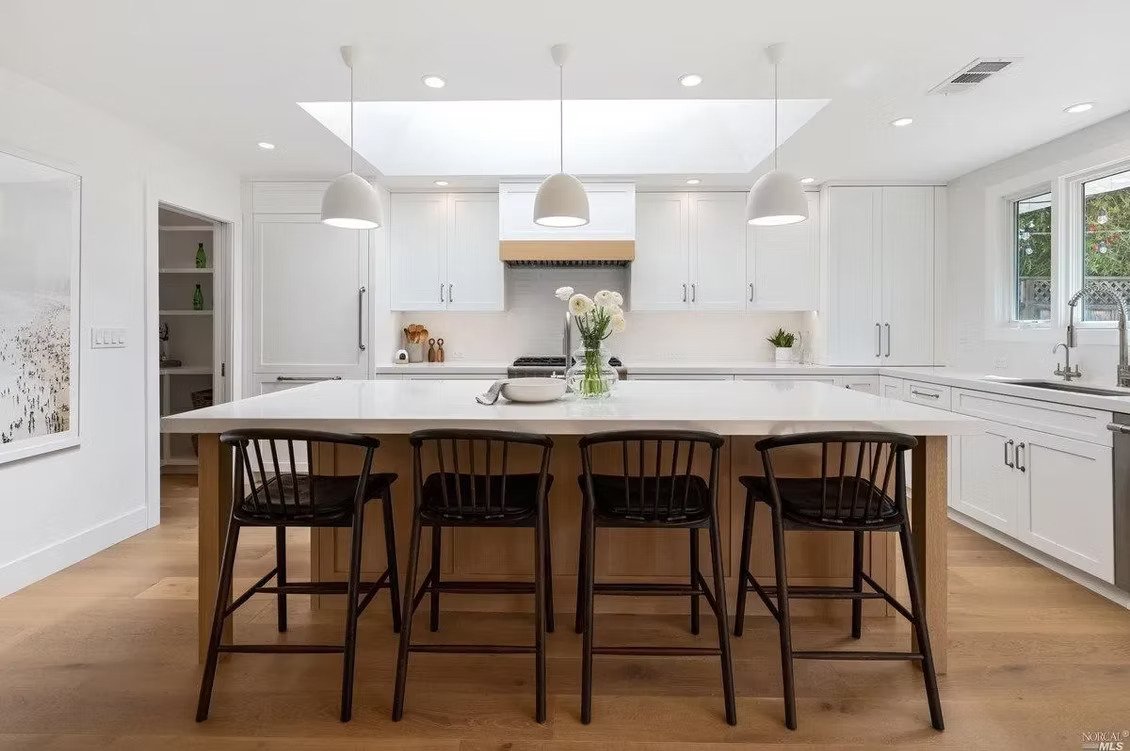 Modern white kitchen with a large island, four black chairs, pendant lighting, and a skylight.