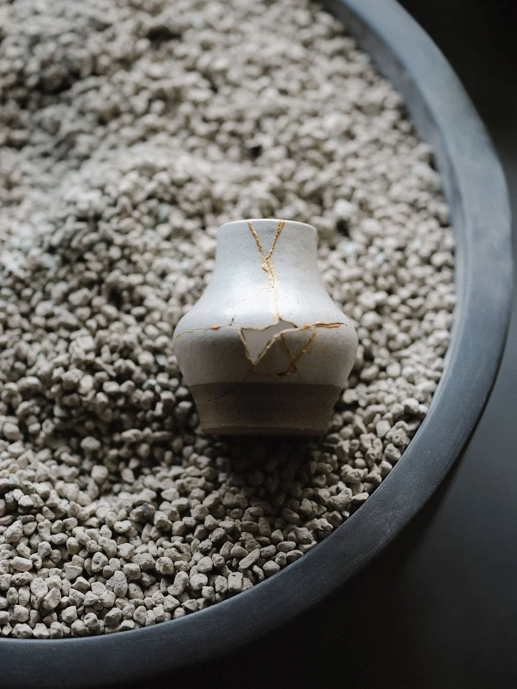 Small white ceramic vase with gold veins on light gray pebbles in a gray container.
