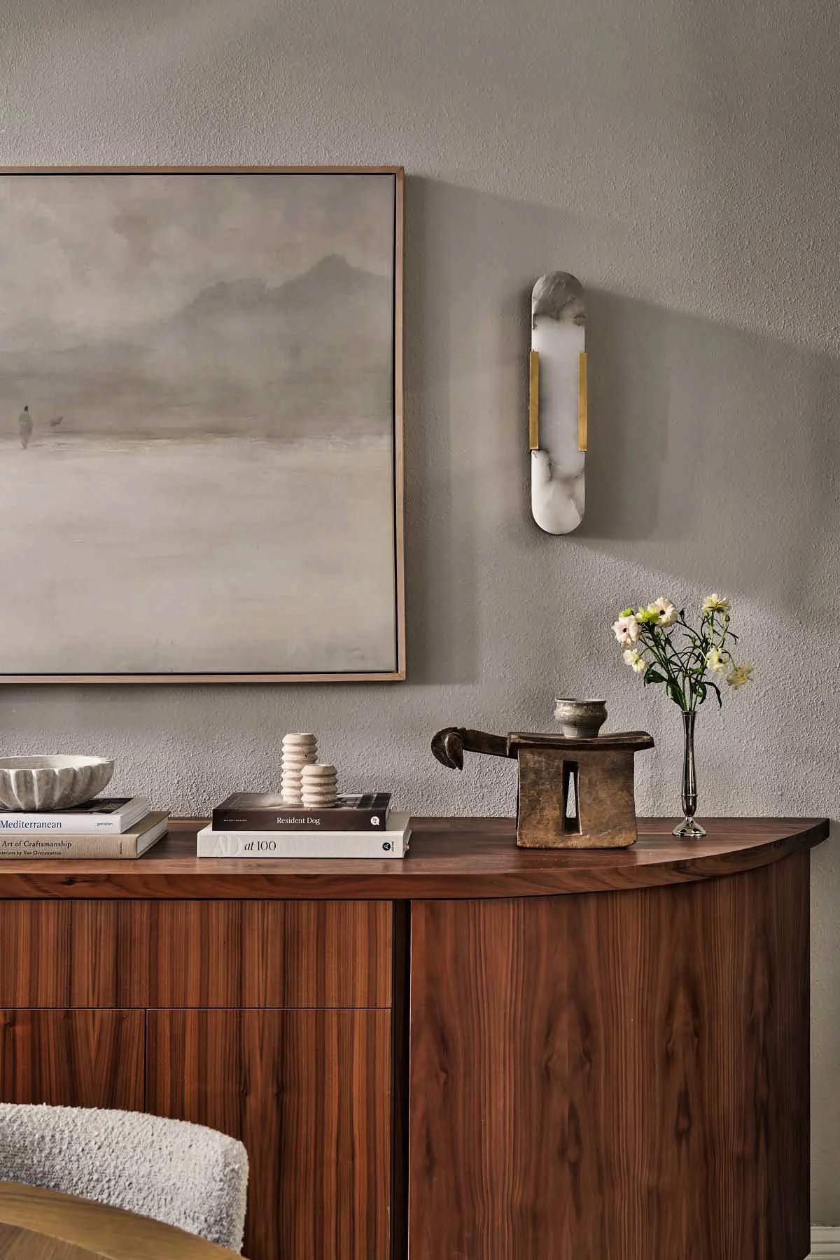 A wooden sideboard with decorative items, including a vase with flowers, books, and small sculptures, against a textured wall with a large watercolor painting and wall-mounted mirror.