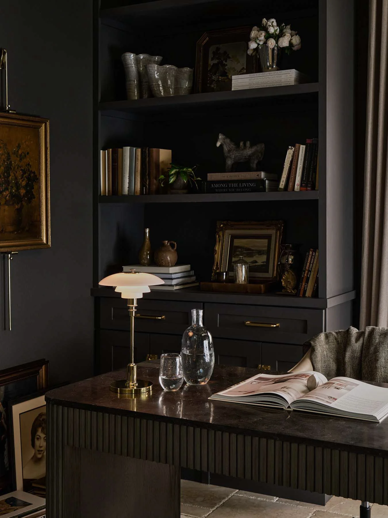A dark-colored bookcase with decorative books, plants, and art objects, alongside a table lamp and a glass water pitcher with glasses on a wooden table, in a dimly lit room with drapes.