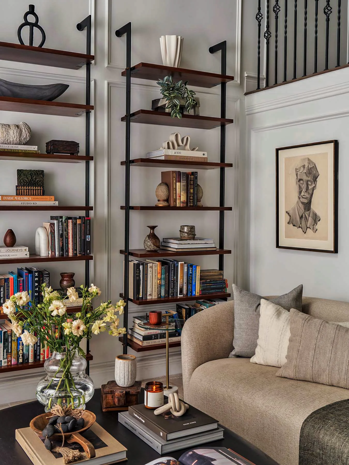 Living room corner with beige sofa and decorative pillows, black coffee table with books, ornaments, and a glass vase of flowers, white wall with framed artwork, gray bookshelf filled with books and decorative objects, staircase railing, and a patter