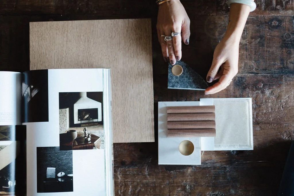 Person arranging samples of wood, stone, and tile on a wooden table, with an open magazine displaying interior design images.