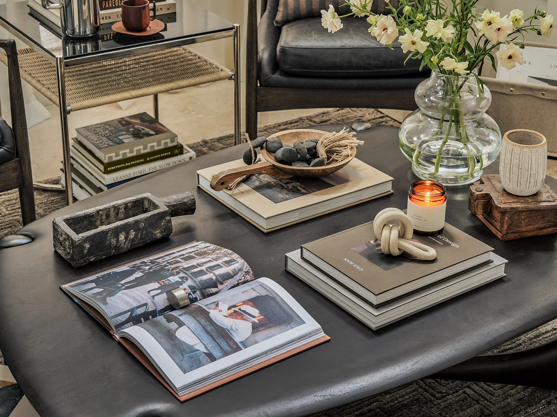 A modern living room table decorated with books, a glass vase with white flowers, a lit candle, and various decorative objects. A black leather chair and a side table with books are visible in the background.