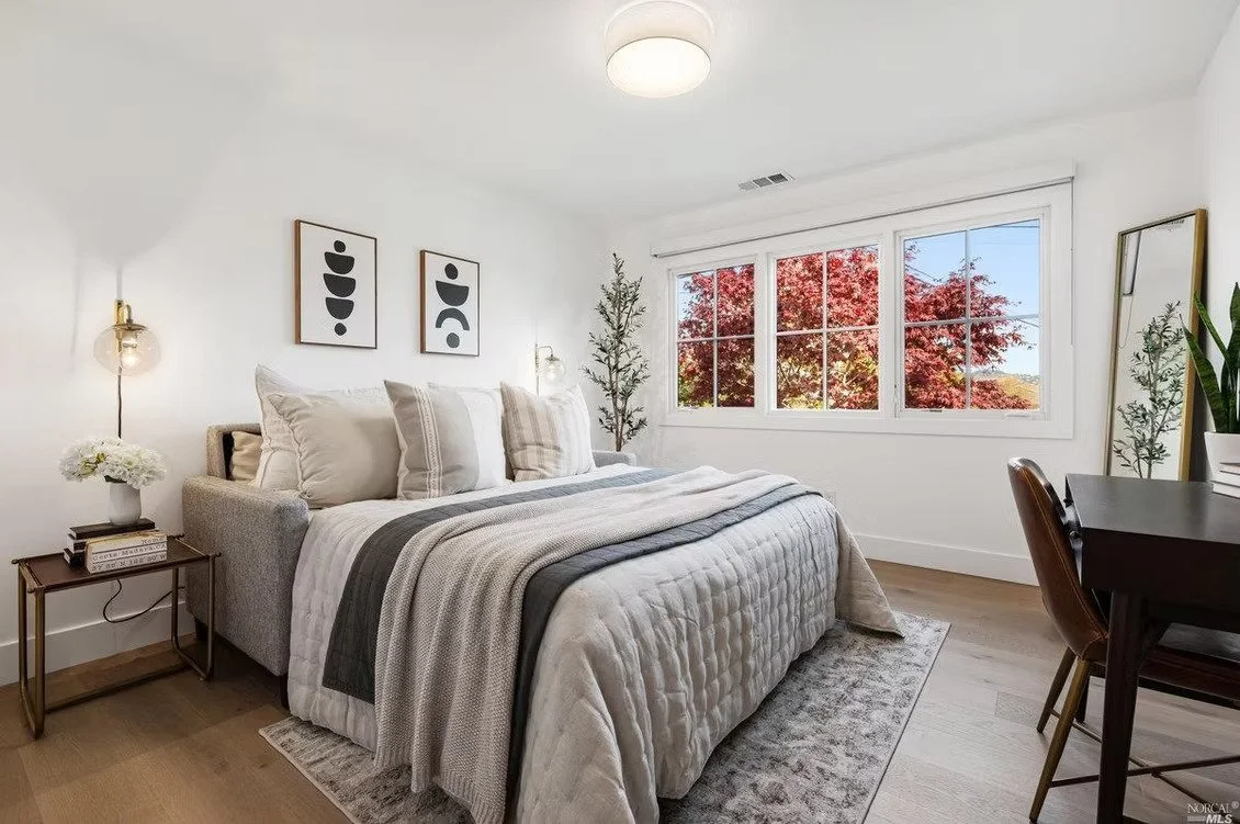 A cozy bedroom with a large bed, white bedding, and decorative pillows. There are two framed abstract art pieces above the bed, a nightstand with a small flower arrangement, and wall-mounted lamps. Large windows show red-leafed trees outside, and a m
