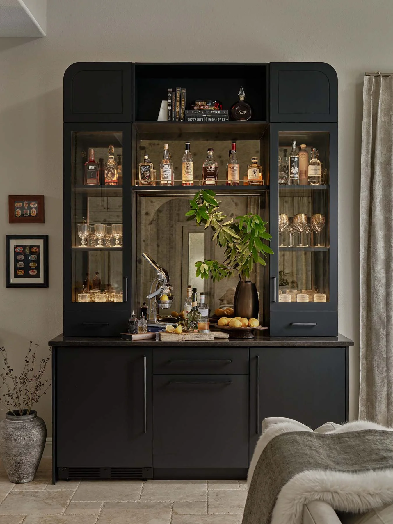 A black bar cabinet with glass doors displaying various liquor bottles and glassware, placed against a neutral-colored wall with framed artwork. A large plant in a vase is on the counter, and part of a beige armchair with a fur throw is visible in th
