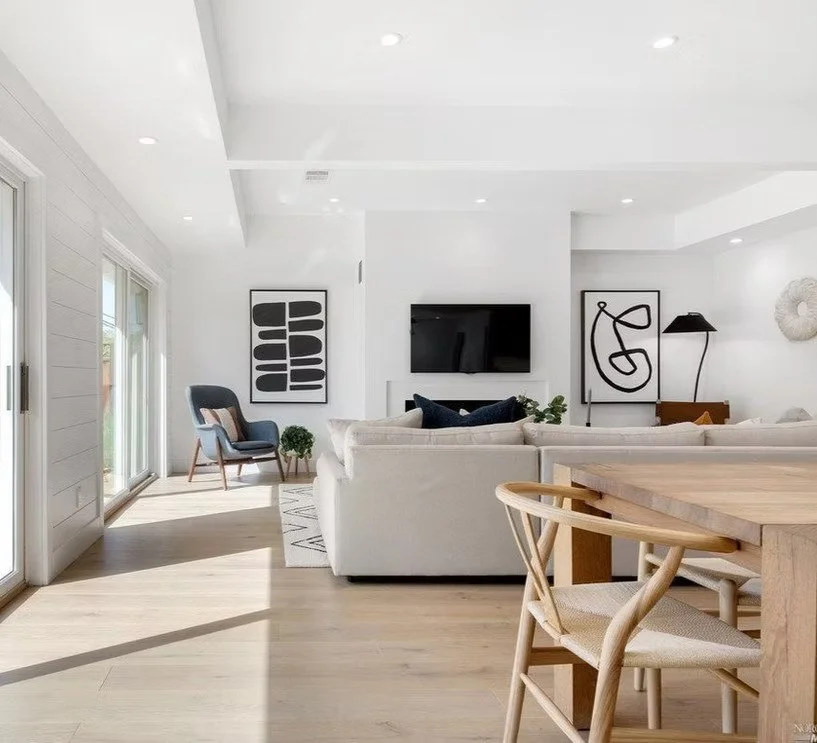 Bright modern living room with white walls, light wood flooring, city view through sliding glass doors, a white sectional sofa, a light wood dining table with chairs, black and white abstract artwork, a wall-mounted TV, a blue armchair, plants, and modern decor.