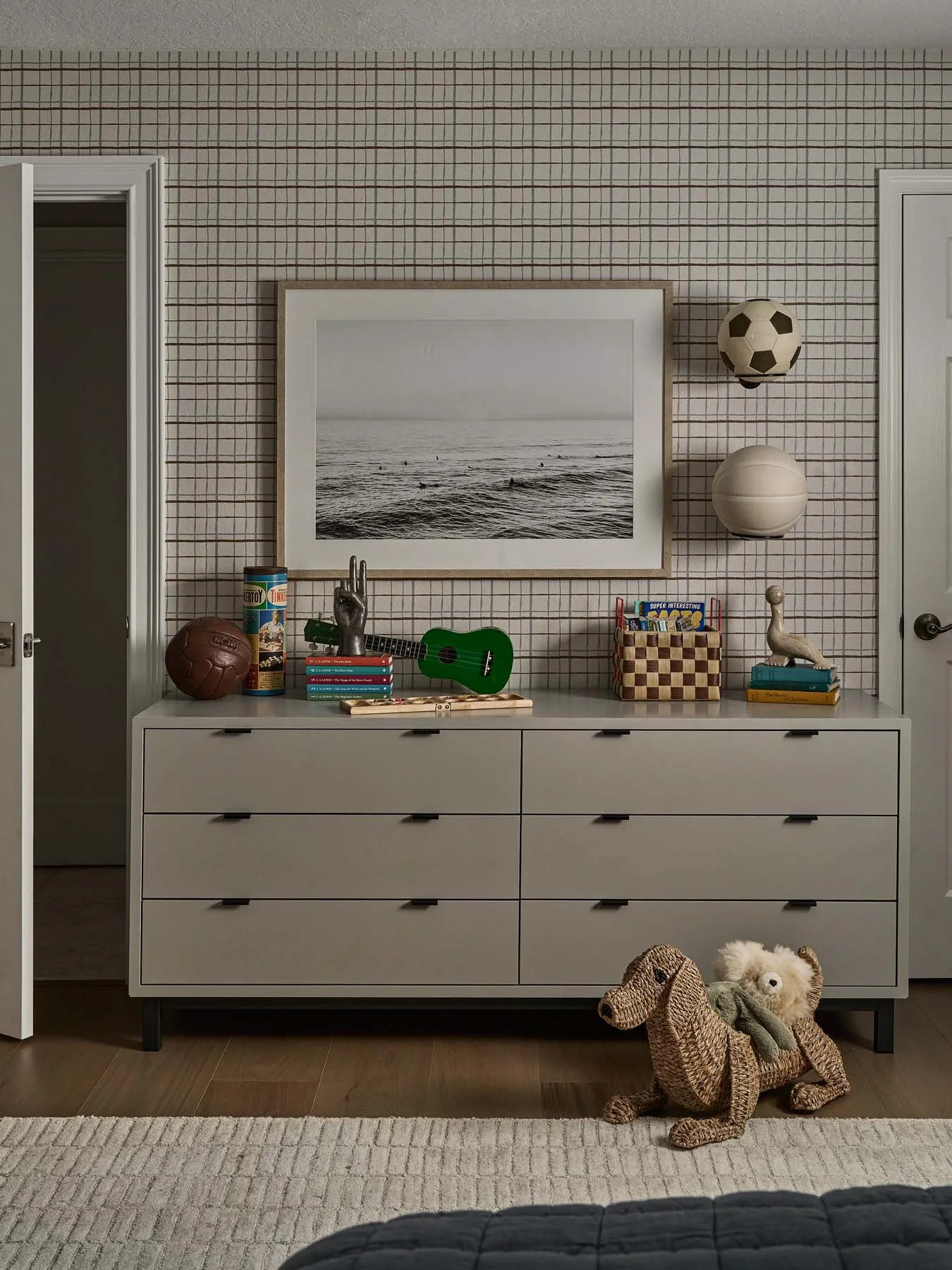 A white dresser with closed drawers in a room with a grid-patterned wall. On top of the dresser are sports items, books, a small figurine, and decorations. There is a framed black-and-white ocean photograph on the wall. Two ball-shaped objects, one r