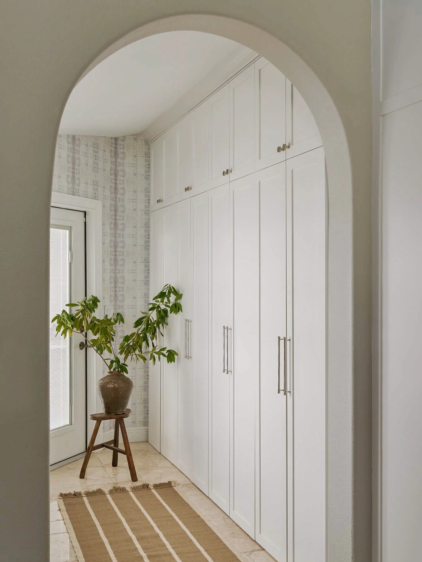 Entryway with tall white storage cabinets, a potted plant on a small wooden stool, a beige and white striped rug, and a door with frosted glass window.