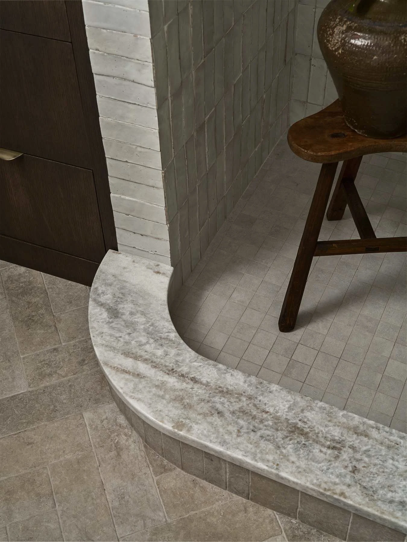 Corner of a bathroom or kitchen with a marble countertop, tiled walls, and a wooden stool with a ceramic pot on top.