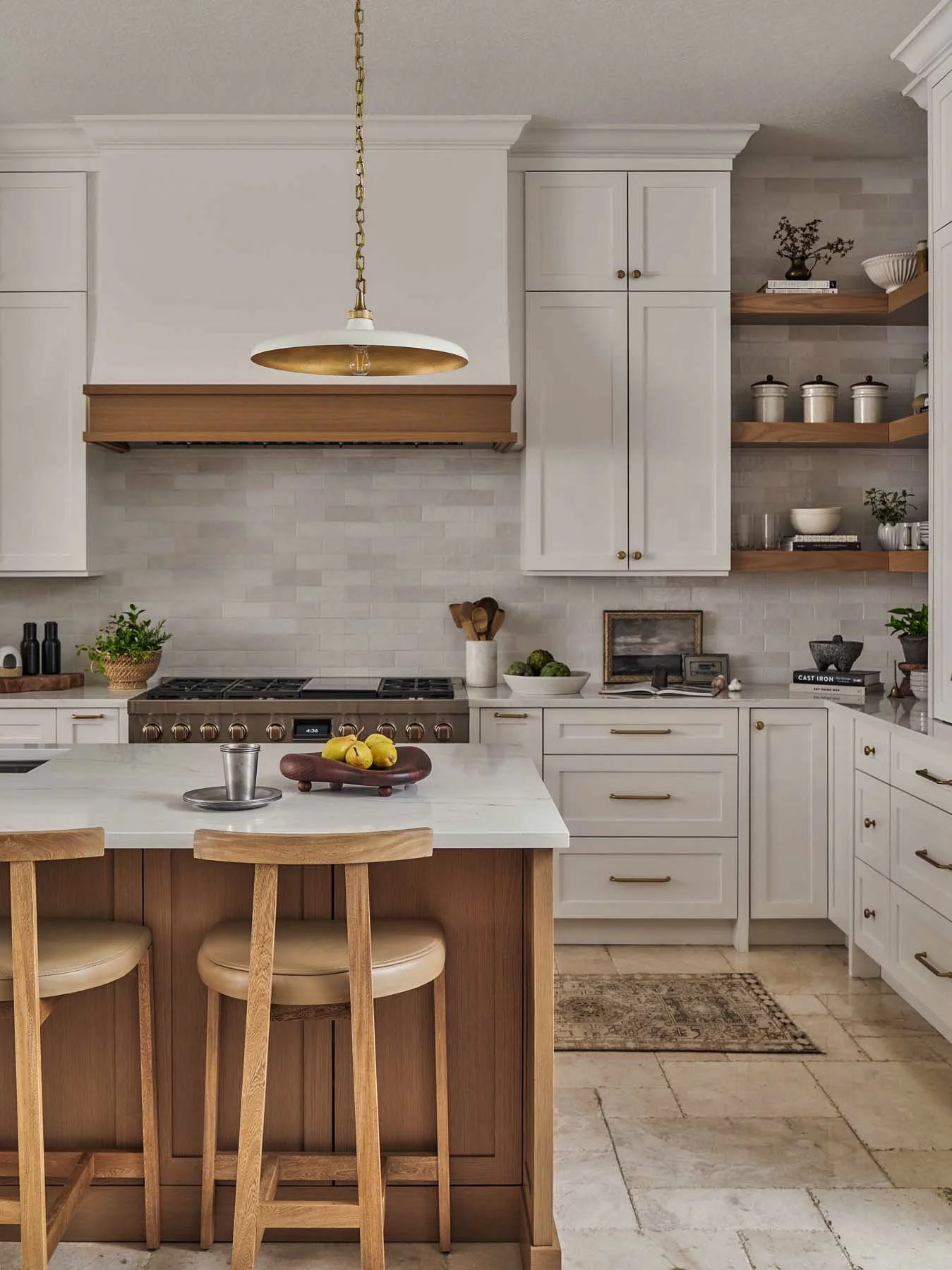 A kitchen with white cabinets, open shelves with bowls and jars, a central island with lemons and a stool, and a stainless steel stove under a wooden range hood, with a brass and white pendant light hanging from the ceiling.