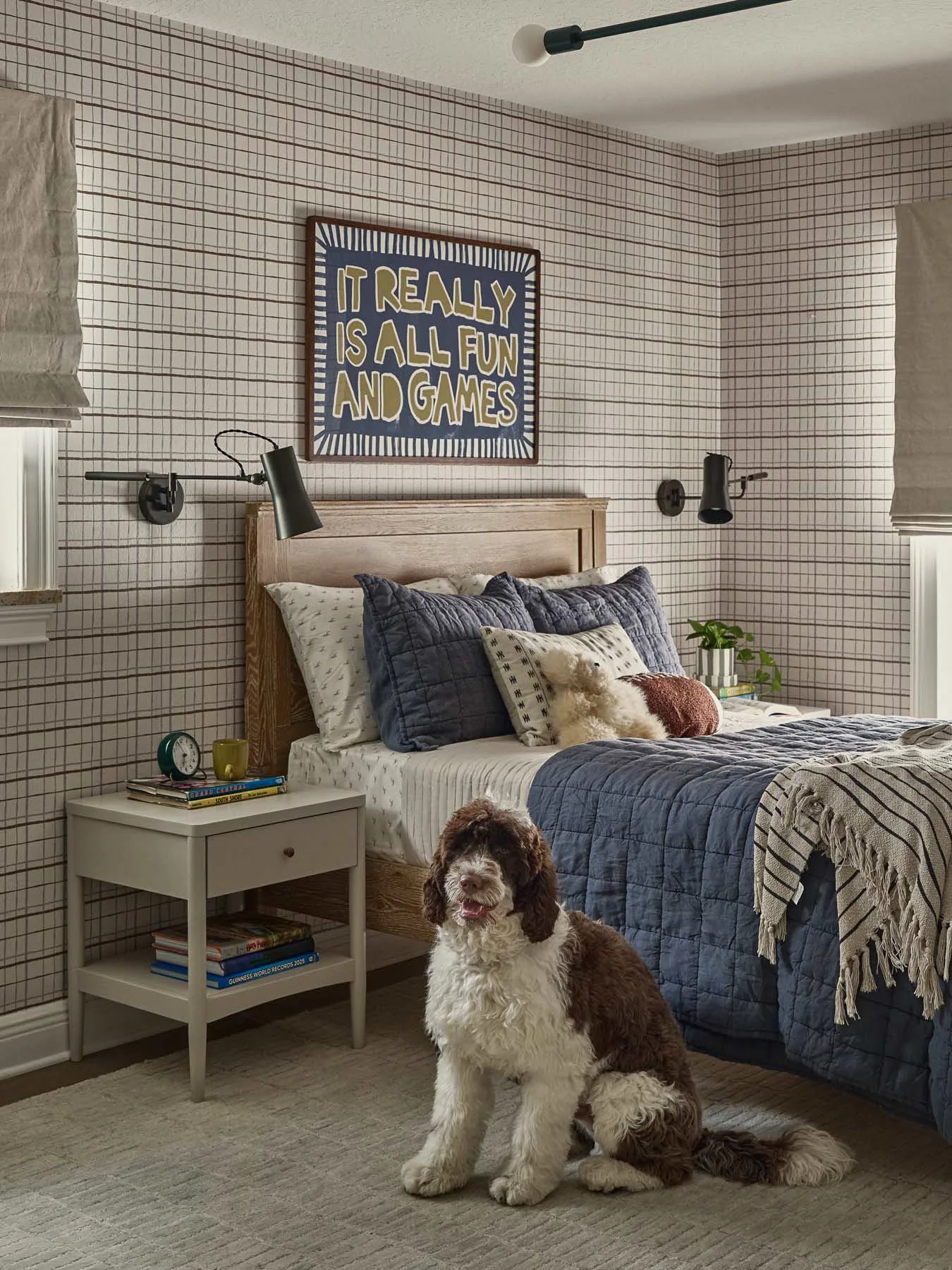 A bedroom with a bed covered in blue and white bedding, decorative pillows, and a blanket. The walls have a checkered pattern, with a framed sign saying 'It really is all fun and games.' There are two black wall-mounted reading lamps on either side o
