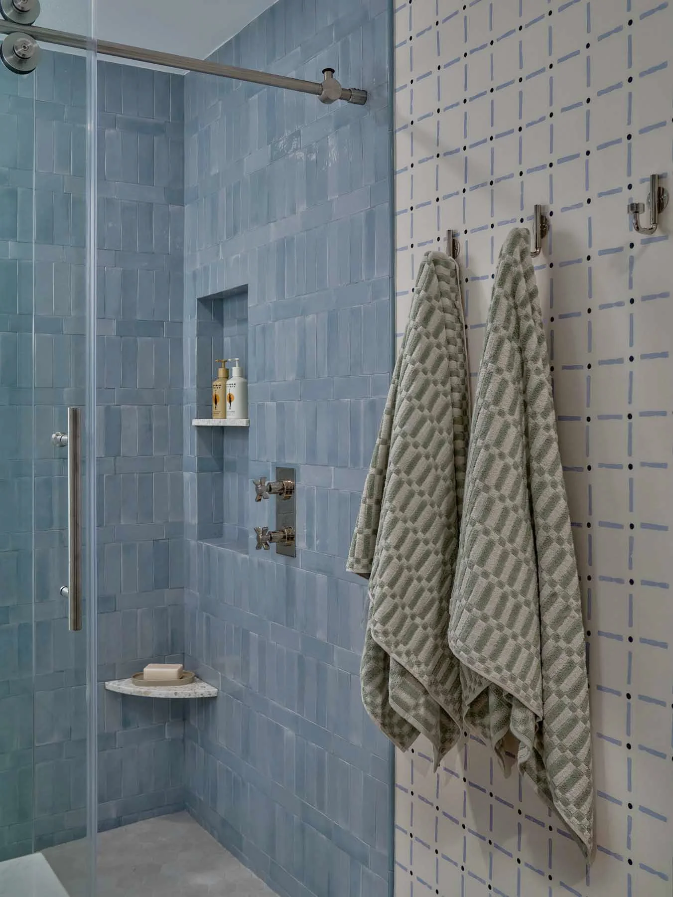 A shower area with light blue tiled walls and a glass door. Inside, there are two bottles of soap or shampoo on a small marble shelf, and a soap dish with soap on the bottom shelf. To the right, there are two striped towels hanging on hooks on a wall