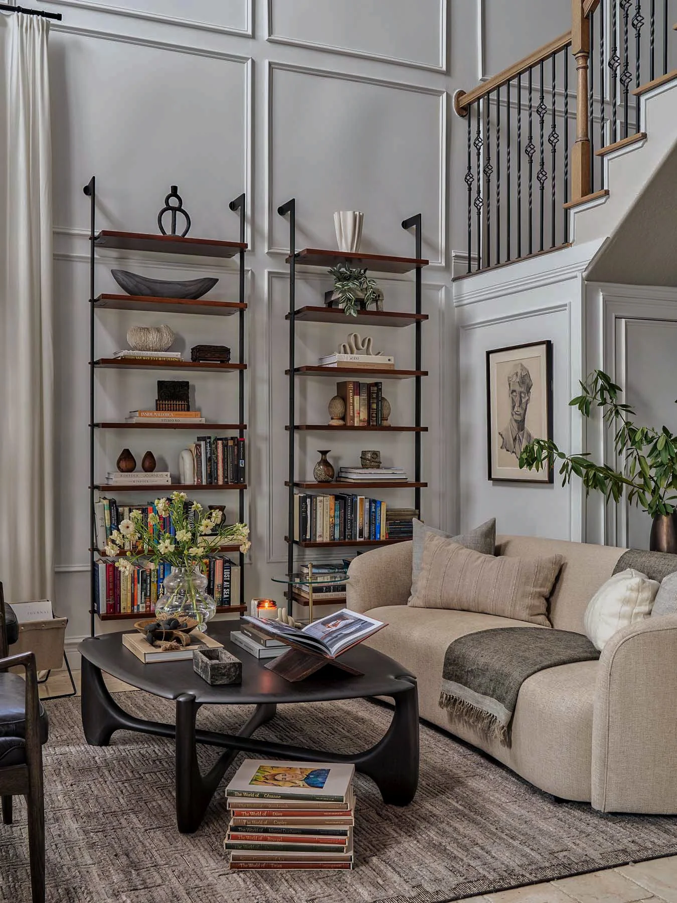 Living room with beige sofa, wooden coffee table, and black shelving with decorative items and books. There is a framed portrait on the wall, and a potted plant near the sofa. The room has high ceilings with white paneled walls and a staircase with b