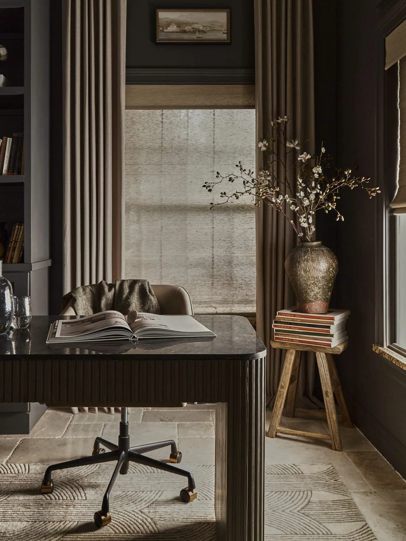 A cozy home office with a marble-top desk, an open magazine, an office chair, a tall ceramic vase with branches of white blossoms, and a small wooden stool with stacked books, all near a window with beige curtains and a painting above.