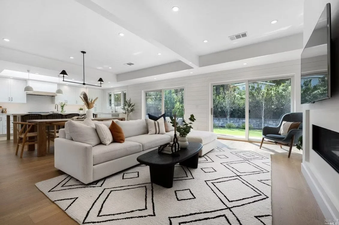 A modern open-concept living room and kitchen with white walls and cabinets, wooden dining table and chairs, a white sofa with pillows, a gray armchair, large glass sliding doors, and a geometric area rug.