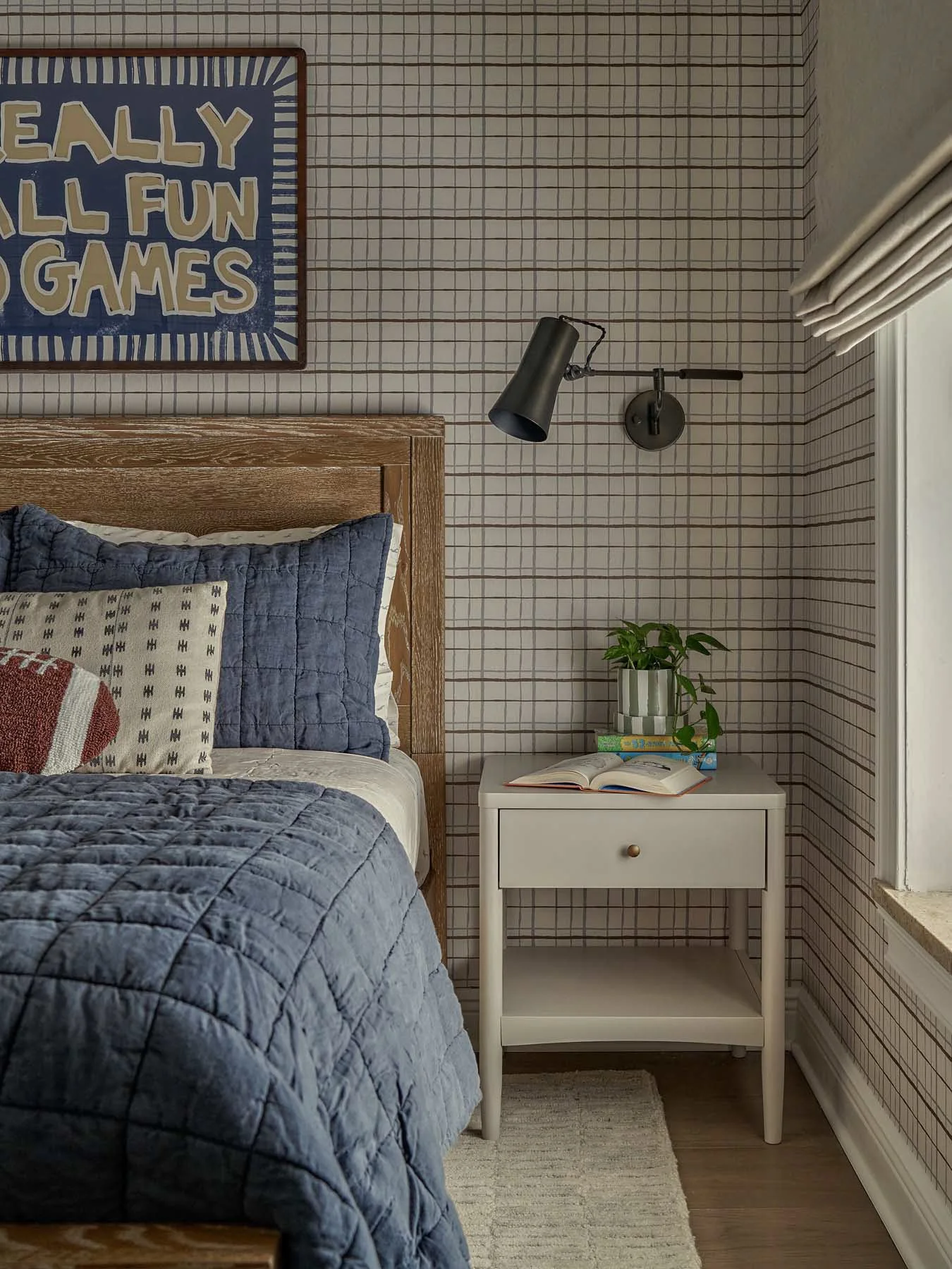 A cozy bedroom corner featuring a bed with a wooden headboard, blue quilted bedding, and pillows. To the right of the bed is a white nightstand with a drawer, an open book, a small stack of books, and a potted plant. The wall has a grid-patterned wal