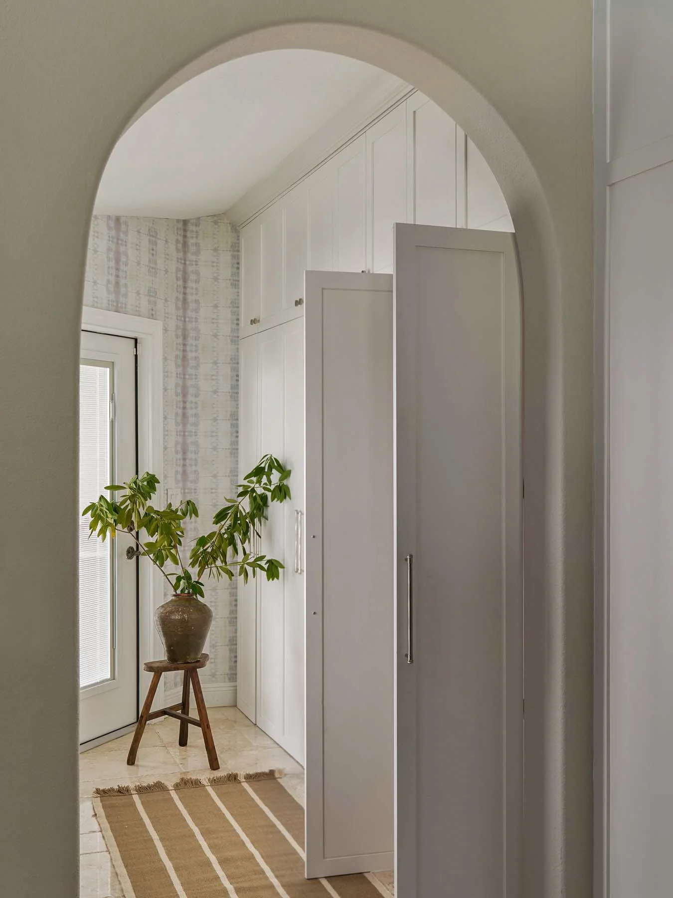 A cozy, well-lit entryway with white built-in cabinets, a small window, a potted plant on a wooden stand, a striped beige and white rug, and a doorway with an arched top.