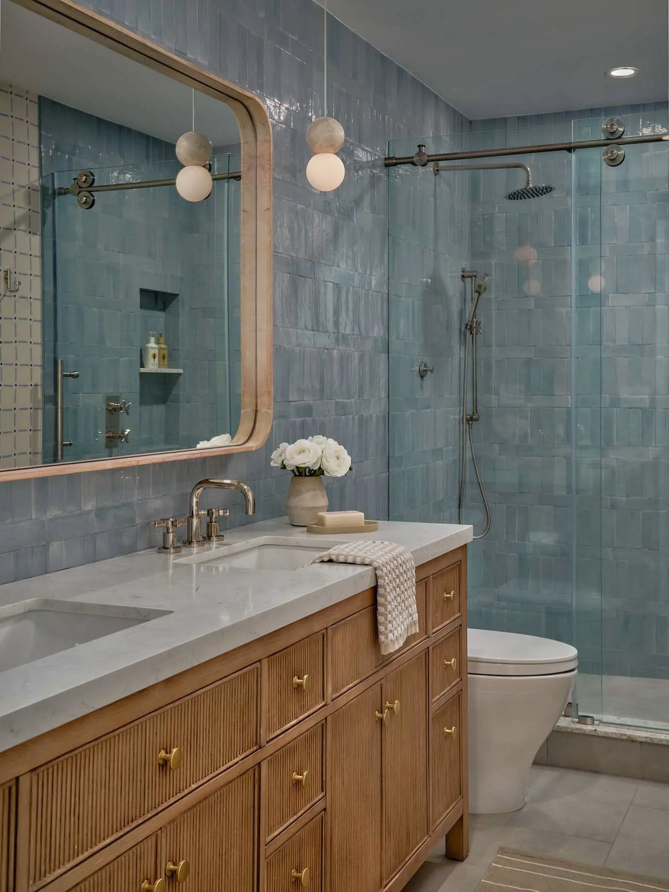 Modern bathroom with light blue tiled walls, a large wooden vanity with a white marble countertop, a mirror, a sink with a brass faucet, and a section of shower area with a rain showerhead and handheld shower. Decor includes a vase of white flowers, 