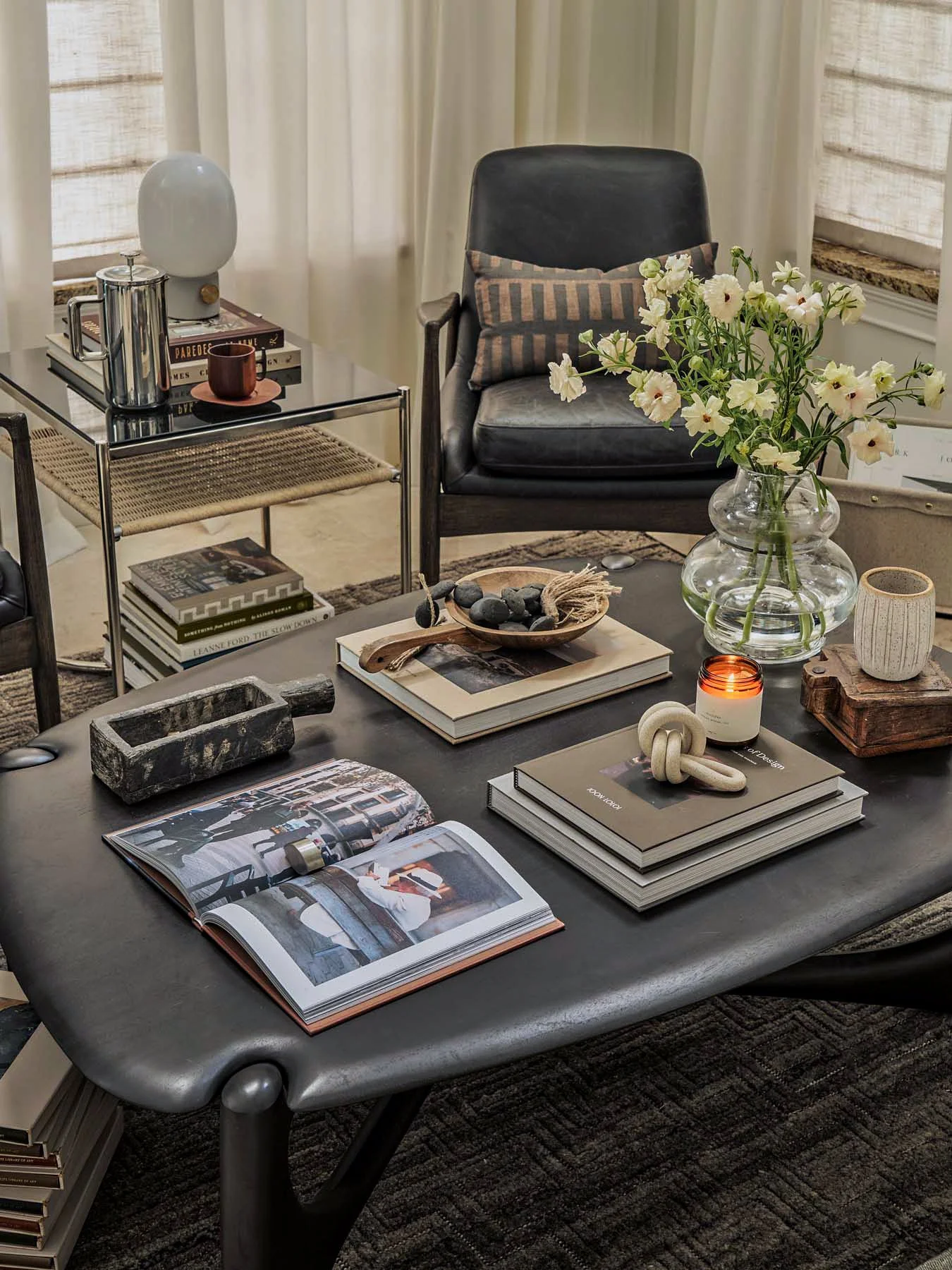 A living room scene with a black leather armchair, a glass and metal side table with books, a table with various books and decorative items, a large glass vase with white flowers, and a dark rug.