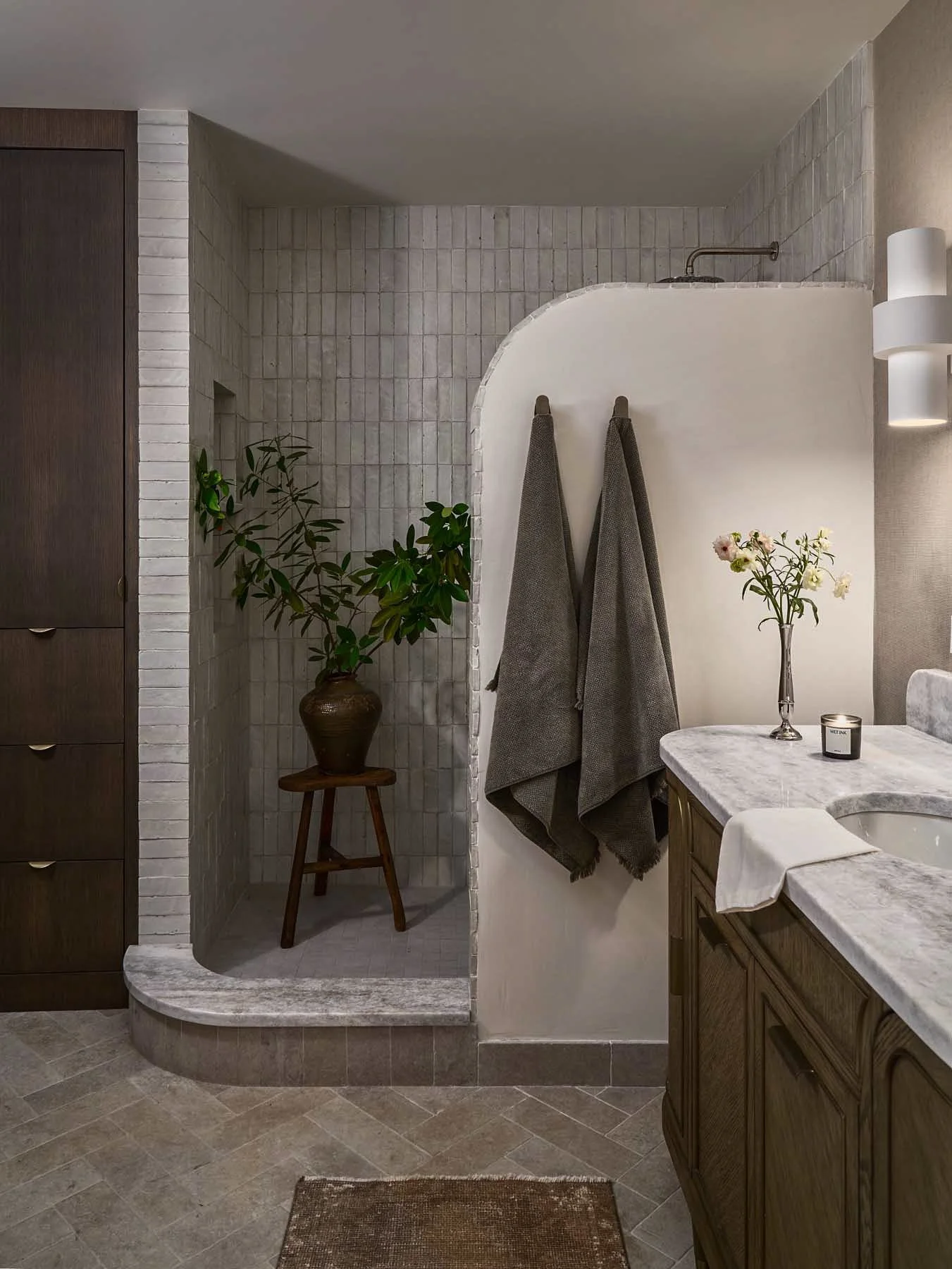 Bathroom with a walk-in shower, gray tiled walls, dark wood cabinetry, and a marble countertop with flowers and a candle. Gray towels hang on a towel rack, and there is a potted plant on a wooden stand.