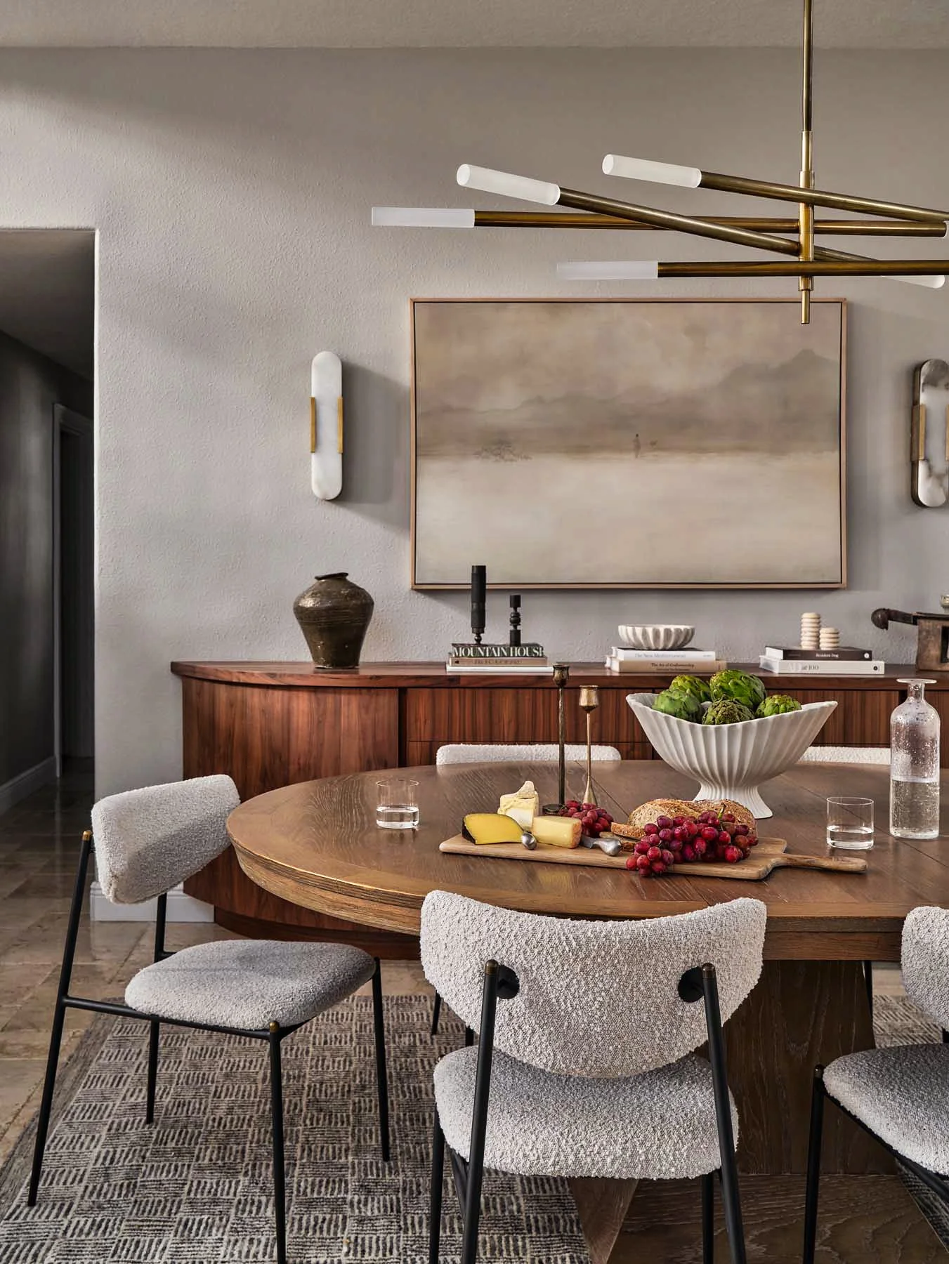 A dining room with a wooden table set with cheese, grapes, bread, and a large white bowl of artichokes. Surrounding the table are four upholstered chairs. In the background, a sideboard holds decorative objects, including a dark vase, books, candles,