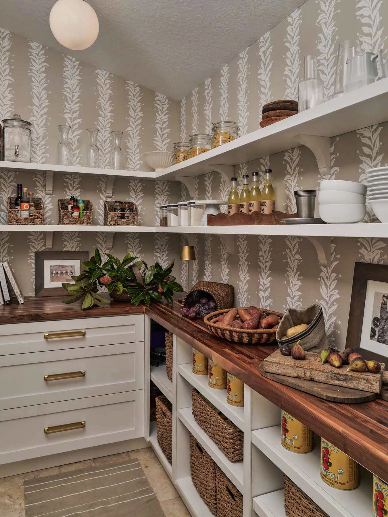 Kitchen pantry with white shelves filled with jars, bottles, bowls, canned goods, baskets, and fresh vegetables. A wooden countertop holds a cutting board with figs and sweet potatoes, with a plant and framed photo nearby.