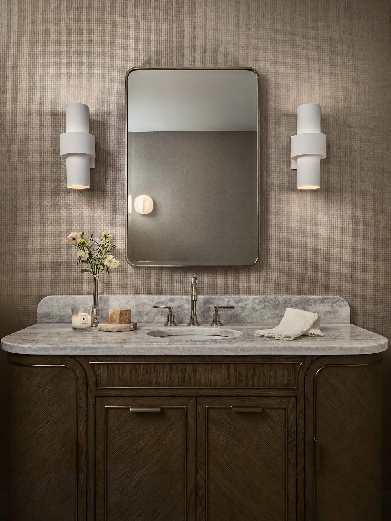 Bathroom vanity with a marble countertop, a mirror, two wall-mounted lights, a small vase with flowers, a candle, a soap dish, and a towel.