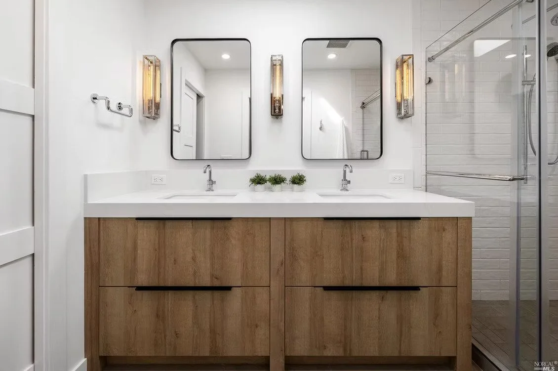 Modern bathroom with double vanity, wood cabinets, white countertop, two mirrors, and a shower with glass door.