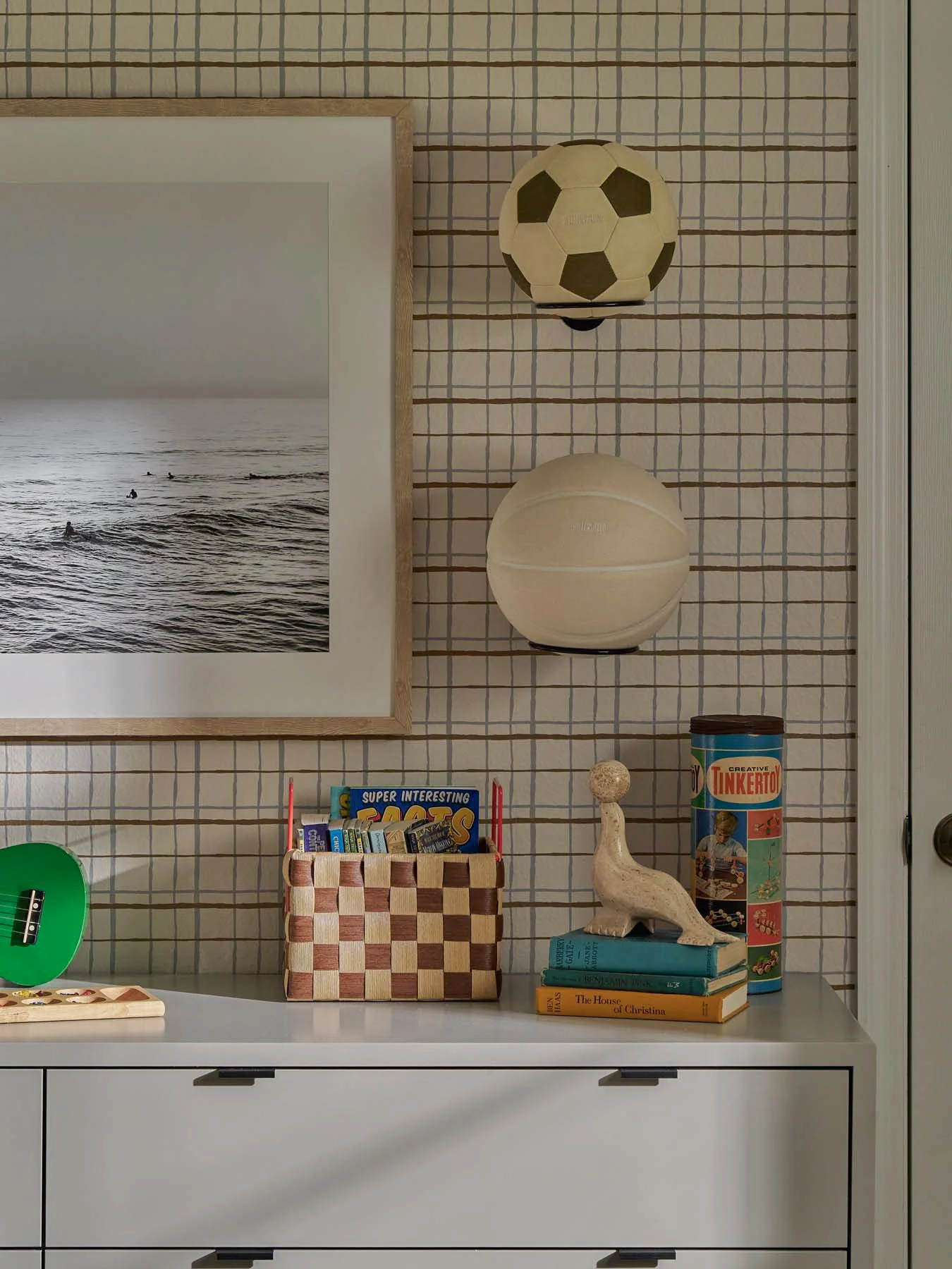 Decorative shelf with a framed black-and-white photograph of ocean waves, a paper lantern designed like a soccer ball, a ceramic bird figurine, books, a cylindrical container labeled "Tinkertoy," and a checkerboard box filled with crayons or markers,