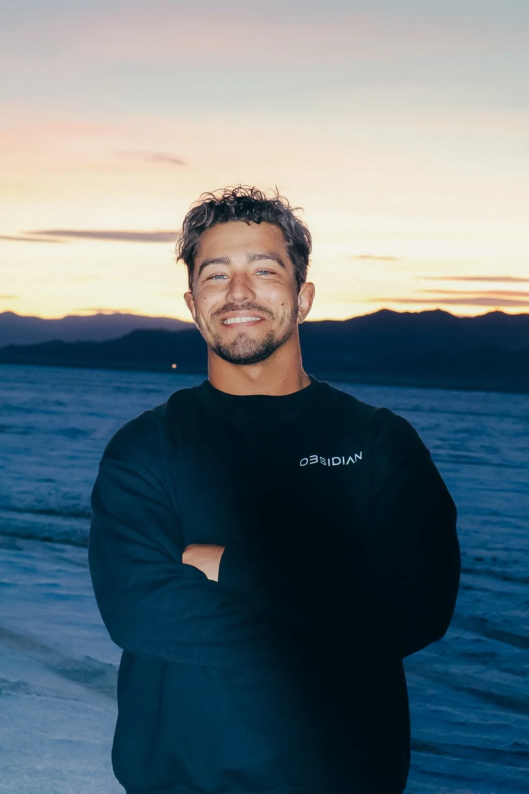 Tavin Stuhr, Orange County personal trainer offering virtual, in-person and hybrid training, smiling in an Obsidian shirt in front of mountains at sunset