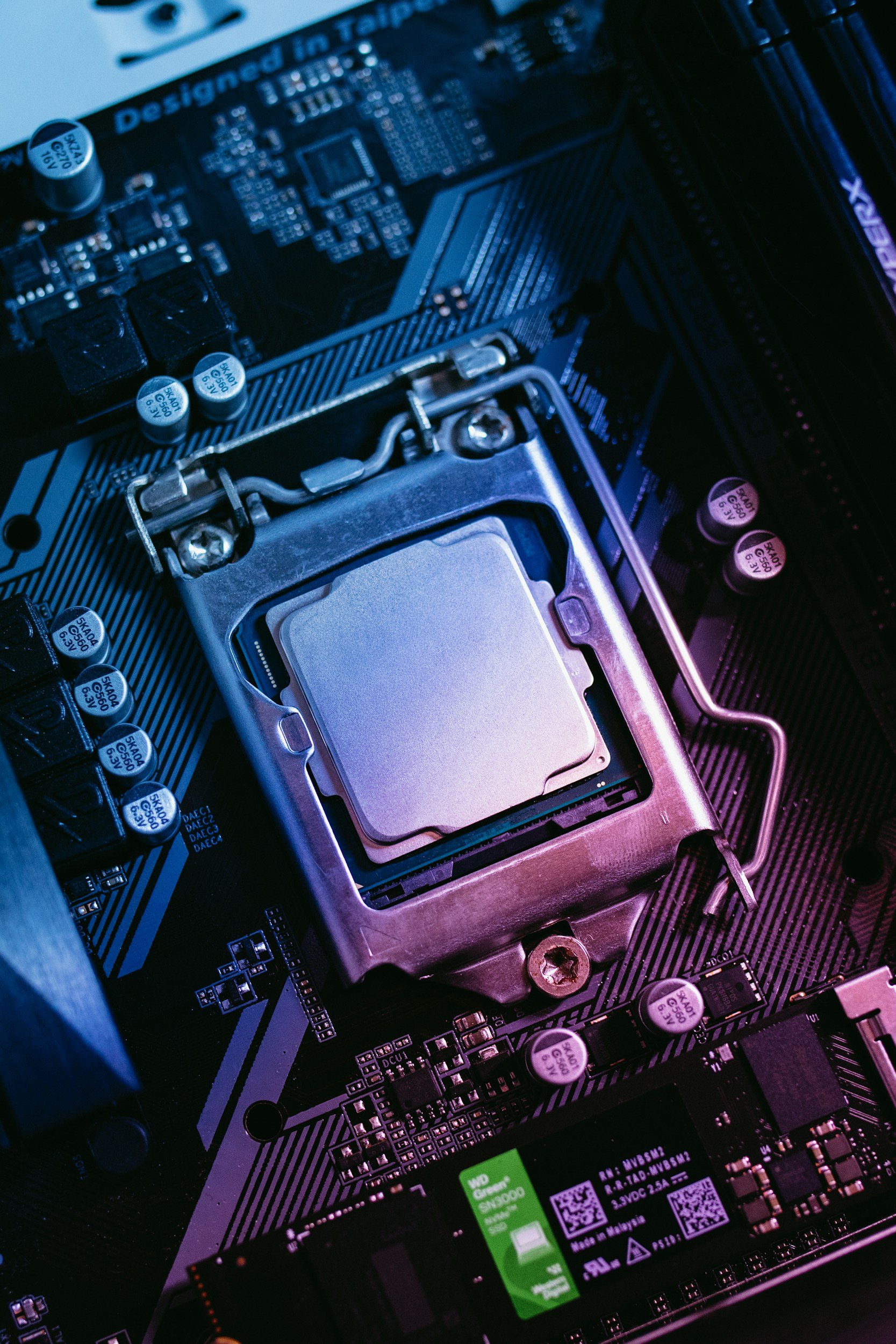 Close-up of a computer motherboard with a CPU socket, capacitors, and various electronic components in blue and pink lighting.