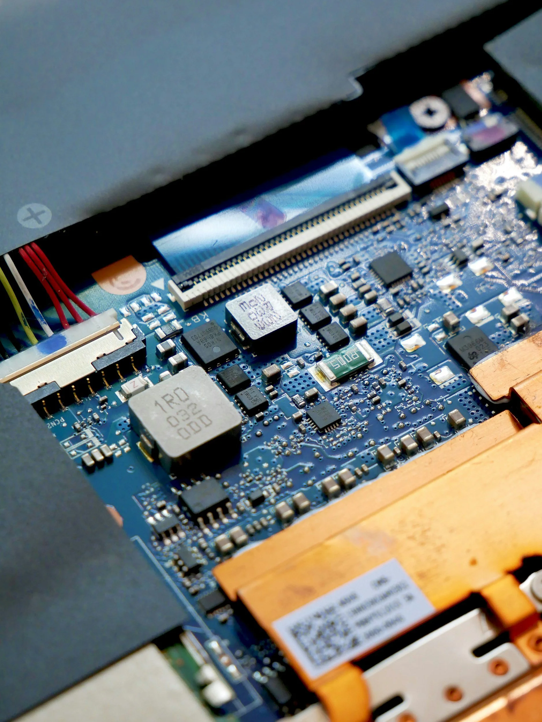 Close-up of a computer motherboard with various electronic components, including capacitors, chips, and a memory module, situated near a laptop's edge.