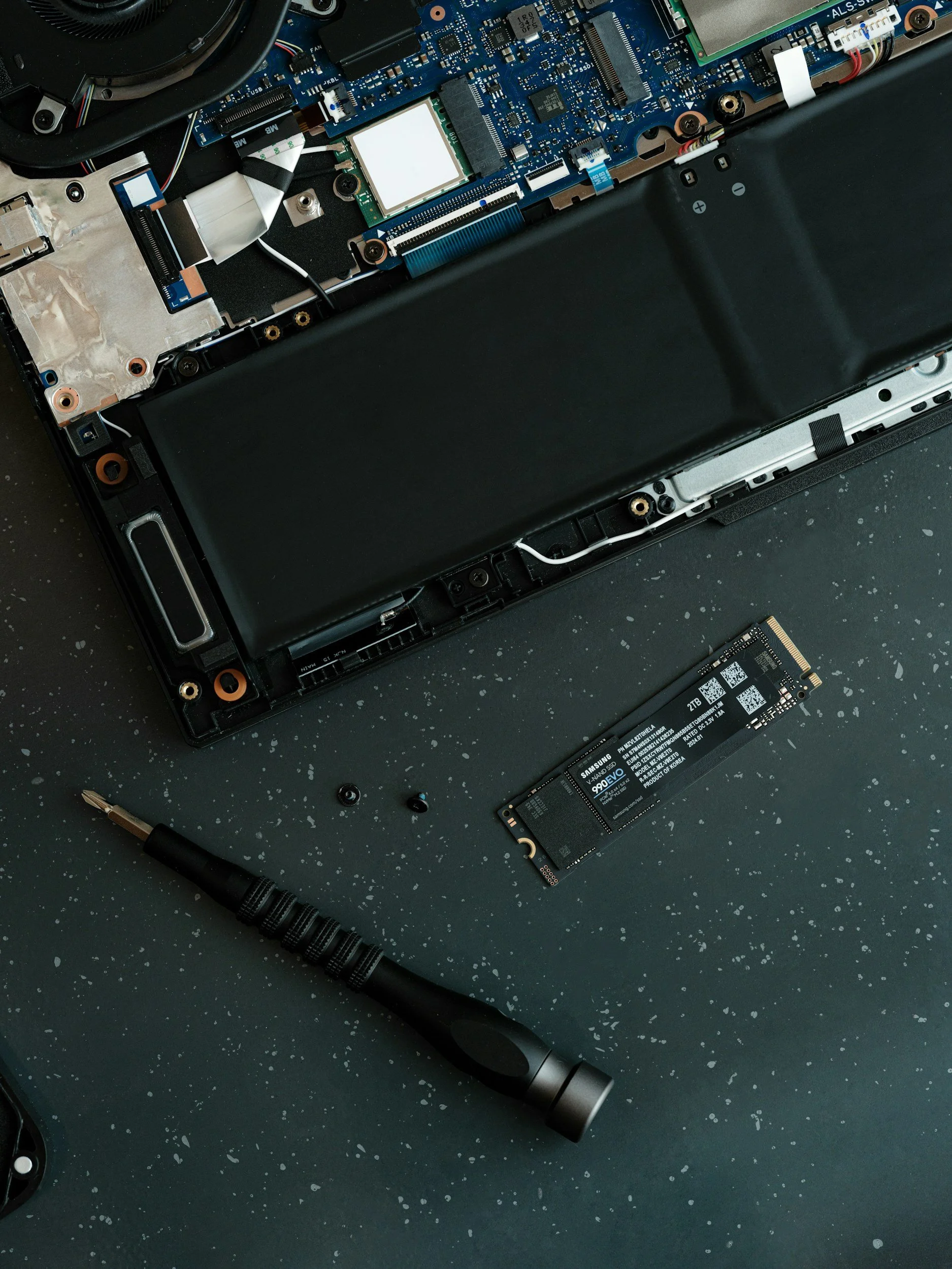 Open laptop with internal components exposed, including a hard drive, various circuit boards, and tools on a dark speckled surface.