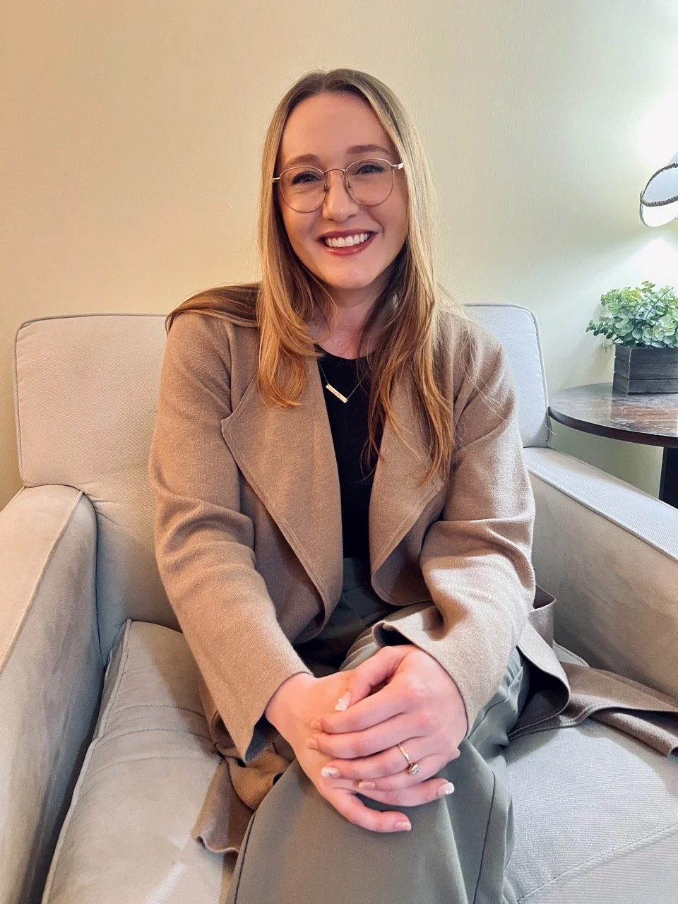 A young woman with glasses and long hair, smiling, sitting on a beige sofa with her hands clasped. She is wearing a tan blazer, a black top, and a ring on her left hand. There is a side table with a green potted plant and a lamp in the background.