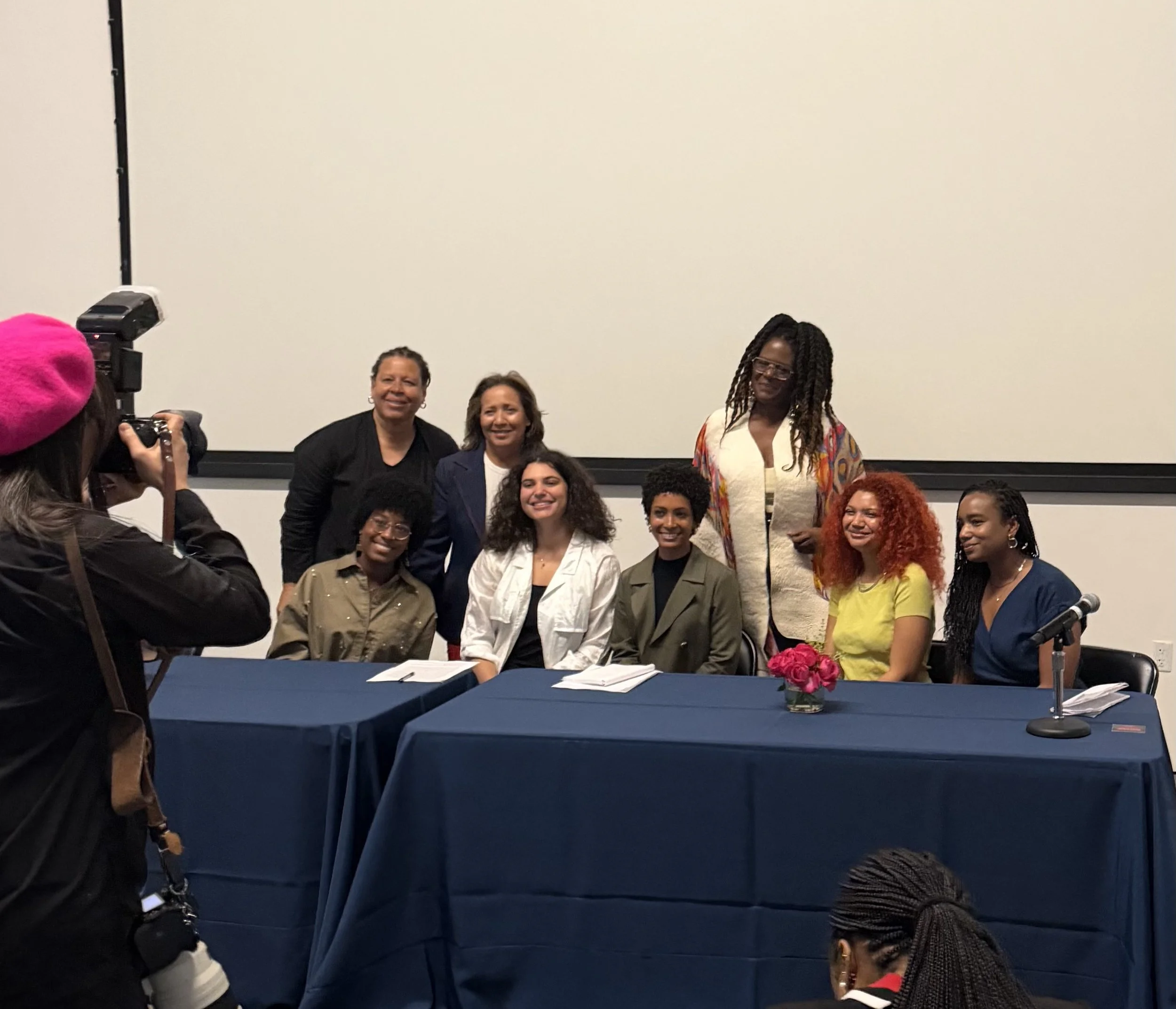 Group posing together in front of a blank white wall, some sitting and some standing, during an indoor event. A table with a blue tablecloth and a vase with pink flowers is in the foreground.