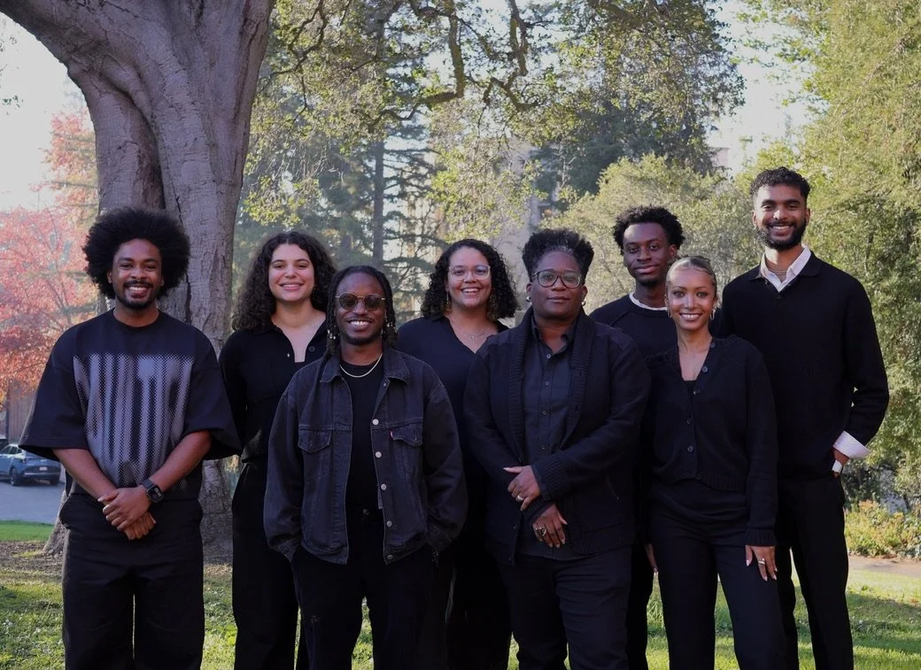 Group of eight people standing outdoors near a large tree with green foliage, smiling at the camera.