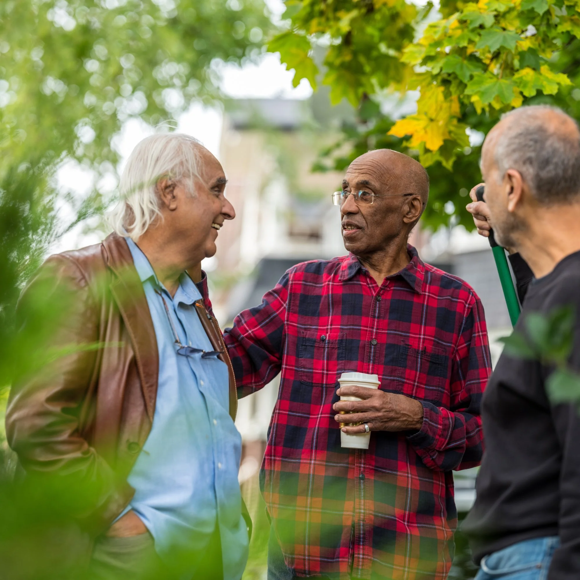 Three elderly men having a conversation outdoors under trees, with one holding a coffee cup, in a park-like setting.