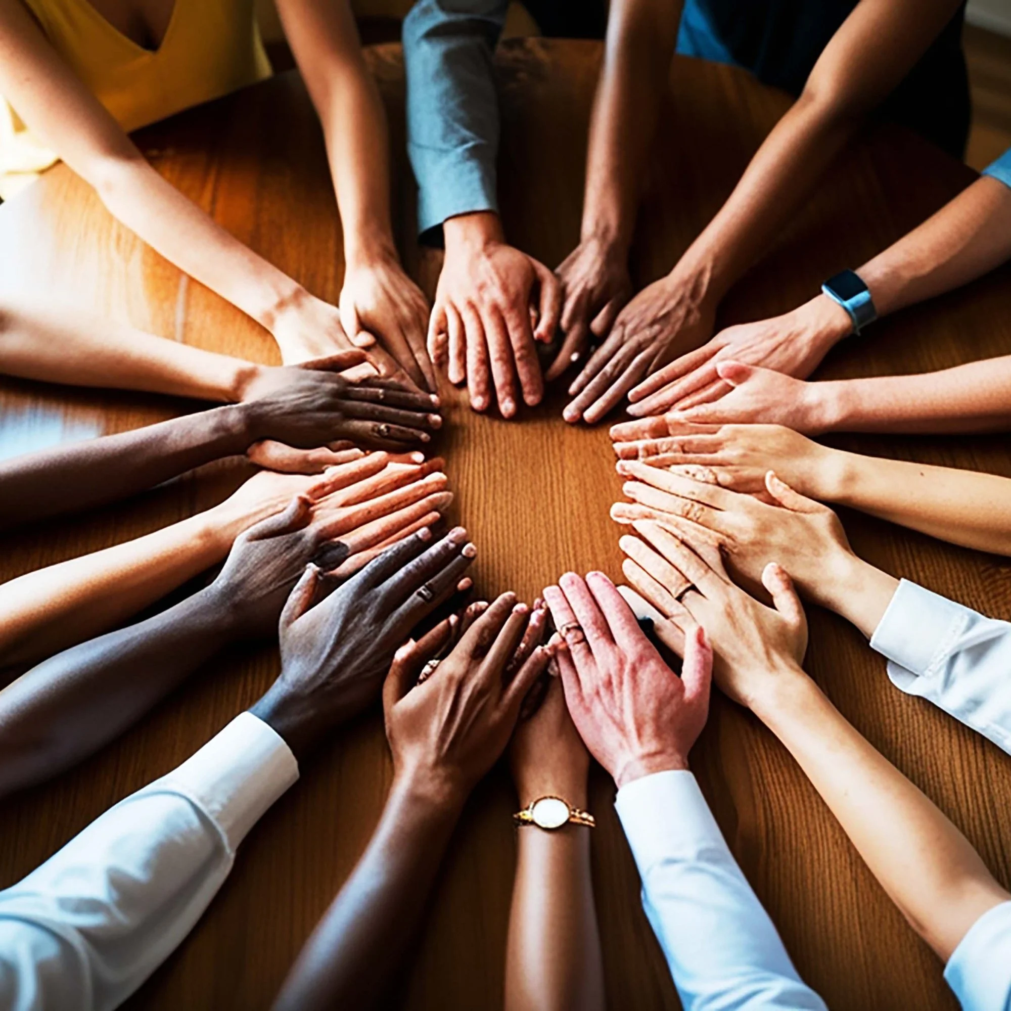 Multiple hands of diverse individuals placed together in the center of a wooden table, symbolizing unity and teamwork.