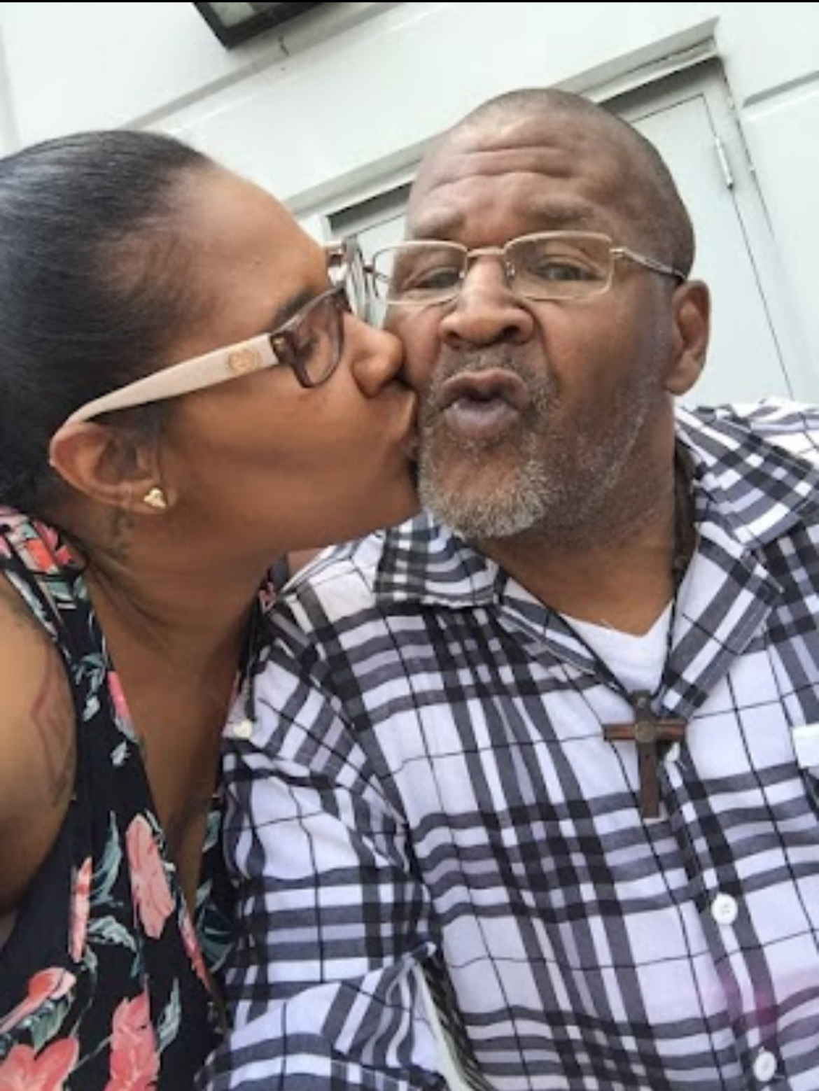 A woman is kissing an older man on the cheek. Both are wearing glasses and smiling, with the woman showing love and affection.