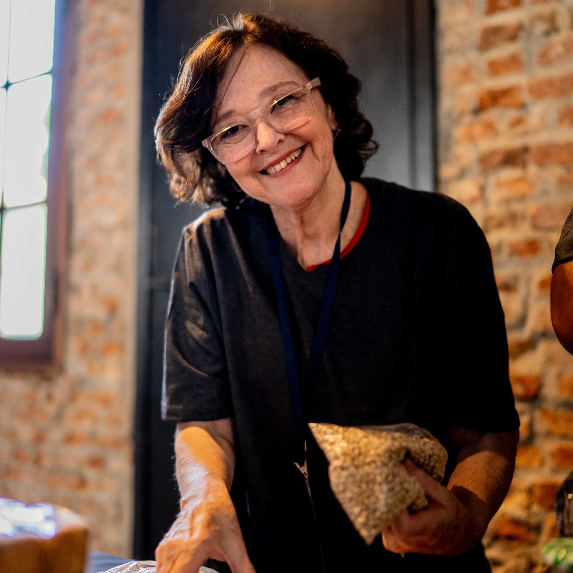 Smiling woman with glasses reaching for something on a table, with a brick wall and a window in the background.
