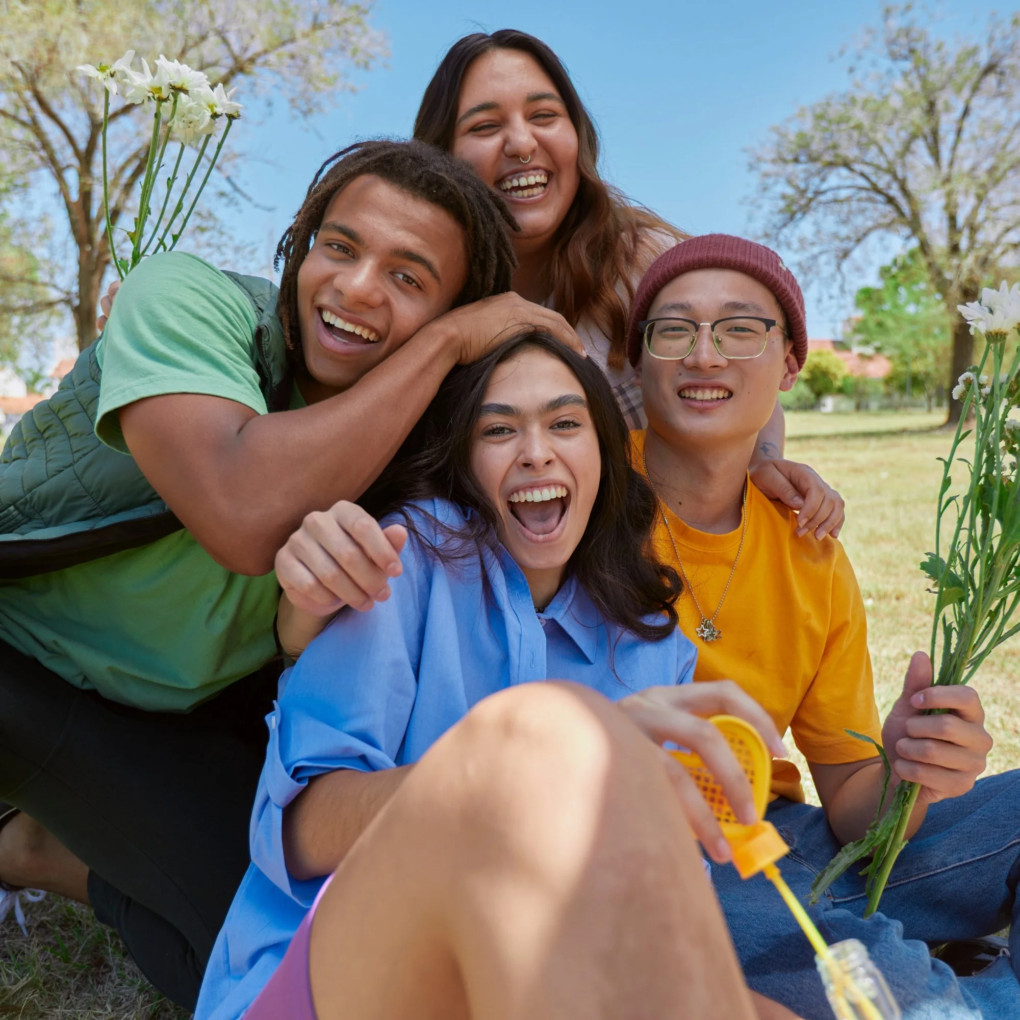 A group of young friends outdoors on a sunny day, smiling and laughing while sitting on the grass among flowers and trees.