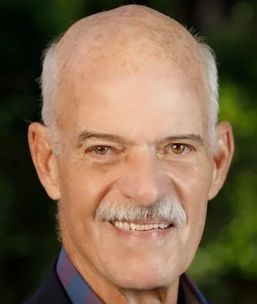 A smiling older man with short gray hair and a mustache, wearing a navy blazer and a checkered shirt, outdoors with a blurred green background.