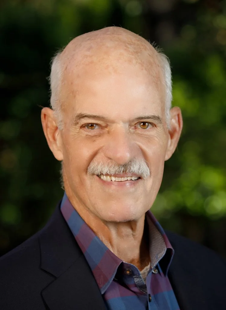 Close-up portrait of an older man with a mustache, wearing a business suit jacket over a plaid shirt, smiling outdoors with a blurred green background.